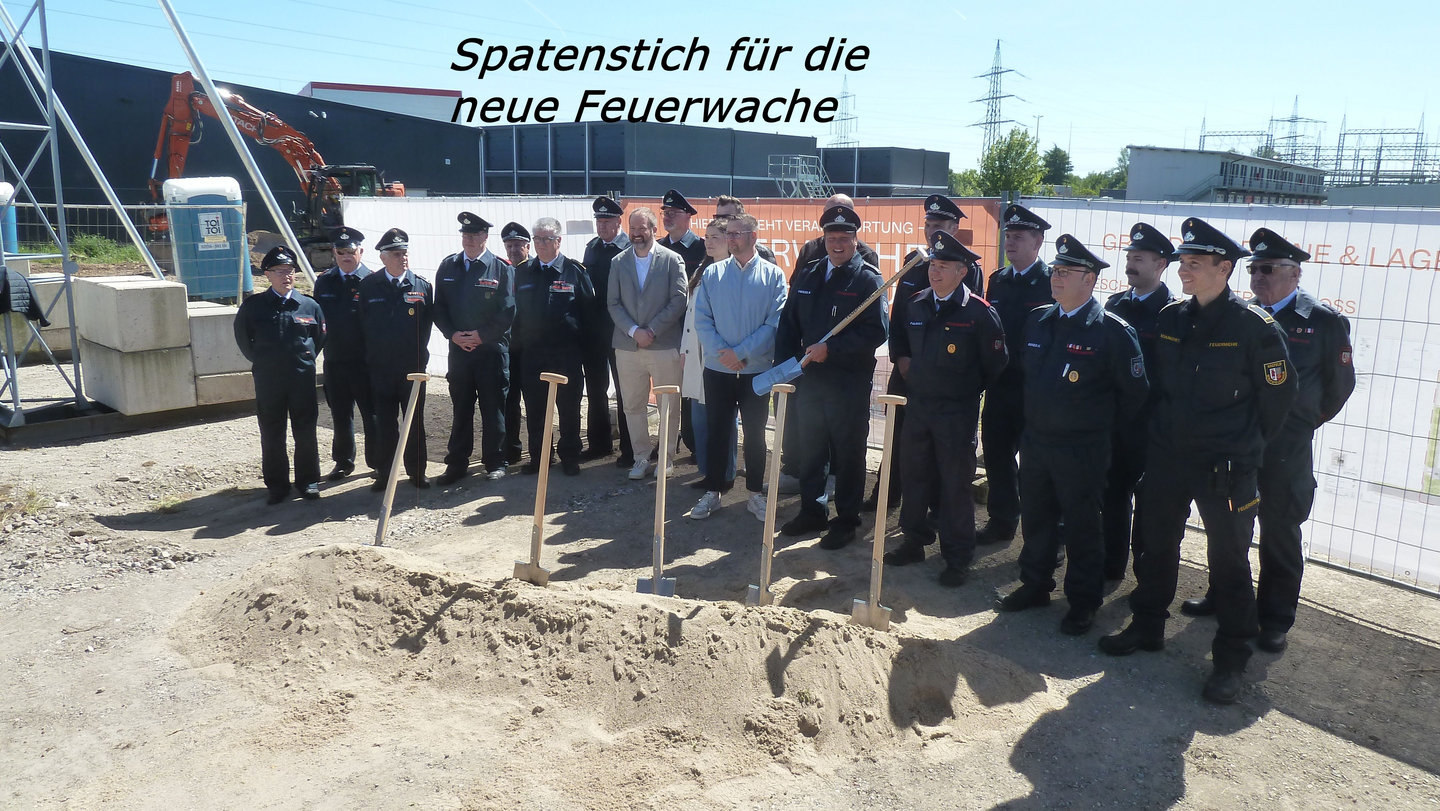 Spatenstich für die neue Feuerwache in Gellep-Stratum, Löschgruppe und städtische Mitarbeiter hinter den Schüppen, vor dem bauzaun, auf dem das bau-Banner zu sehen ist.