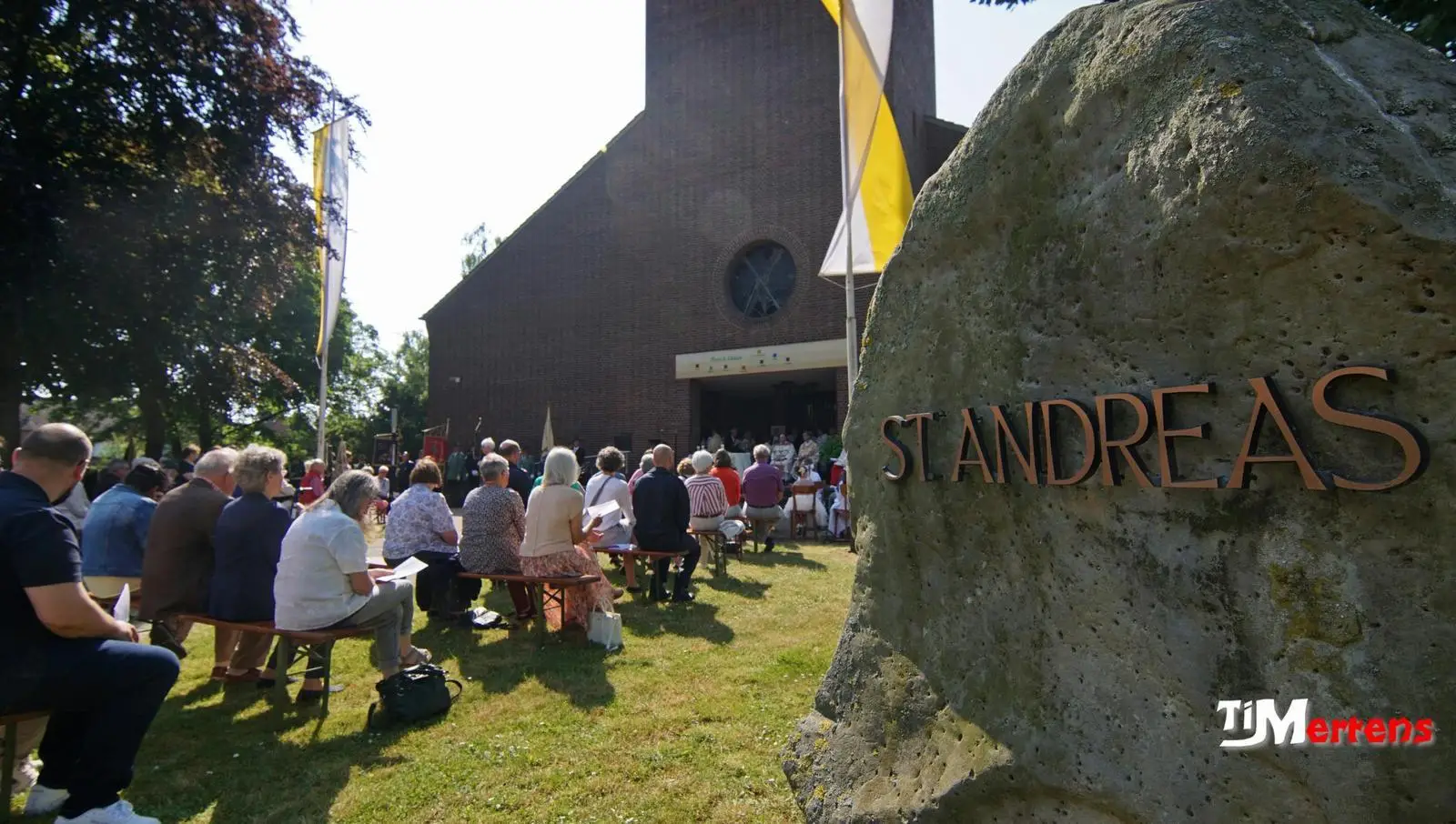 08.06.2023: Foto von Fronleichnam bei heiliger Messe, Sicht auf die Kirche, Stein mit Schiftzug St. Andreas und die Kirchenbesucher auf Bänken.